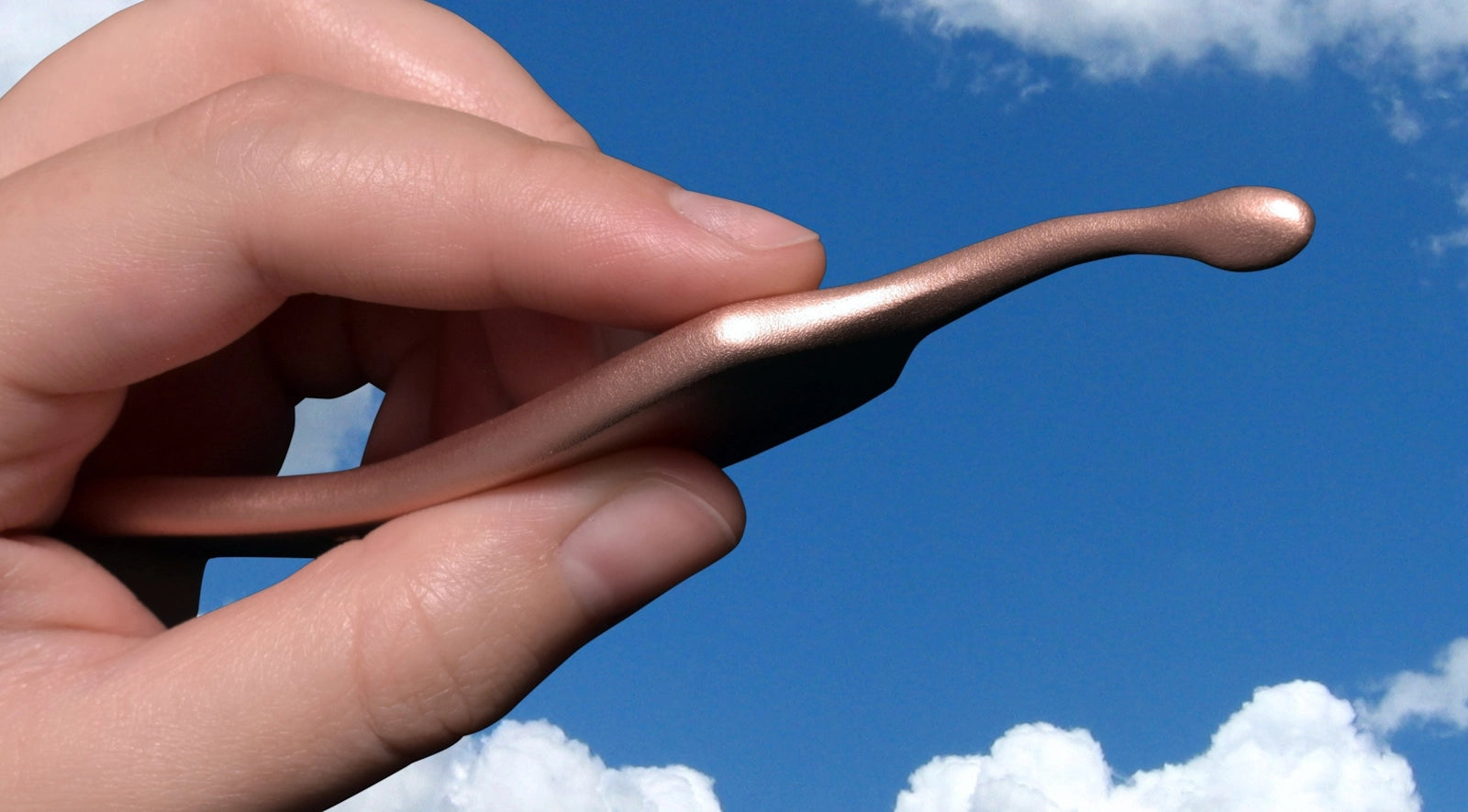 Hand holding a curved, metallic object against a blue sky with clouds.